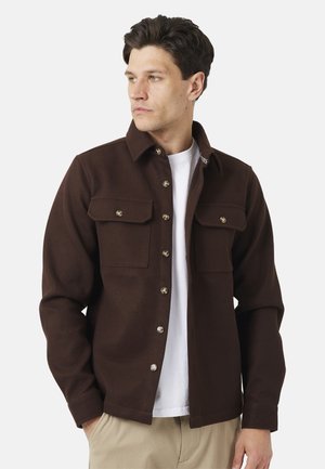 Giovane uomo che indossa una giacca marrone scuro abbottonata sopra una camicia bianca e pantaloni beige, che guarda leggermente verso sinistra su uno sfondo semplice.