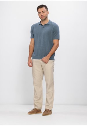 Moški stoji, oblečen v modro polo majico s kratkimi rokavi, bež hlače in rjave priložnostne čevlje, na preprostem belem ozadju.