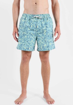 Homme portant un short de bain bleu clair avec des motifs abstraits jaunes et bleu foncé, debout pieds nus sur fond blanc.