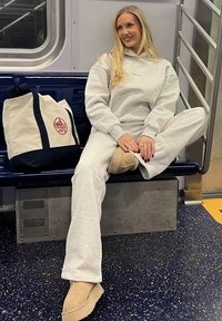 Ung kvinne i grå joggebukse og hettegenser sitter med korslagte ben på en t-bane-benken ved siden av en Trader Joe's handlenett, smilende og ser mot høyre.