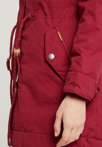 Veste en nylon rouge avec extérieur texturé, poche zippée à fermeture à pression, et cordon de serrage à la taille avec des accents à pois.