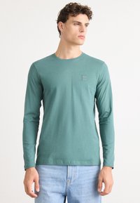Maglietta a maniche lunghe in cotone turchese, collo rotondo, stile aderente. Presenta un piccolo logo quadrato sul petto, abbinata a jeans azzurri.