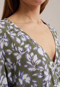 Top floral din material verde măsliniu, cu decolteu în V, având modele de flori albe și lavandă, textură fină și croială lejeră.