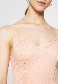 Camisole en dentelle pêche avec de fines bretelles, présentant des motifs floraux délicats et une texture lisse, conçue avec un décolleté en forme de scoop.