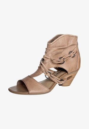 Beżowy skórzany sandał z niskim koturnem, z paskami krzyżującymi się z metalowymi pierścieniami i zaokrąglonym, otwartym noskiem.