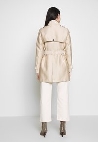 Béžový trench coat s páskem v pase, dvoumřížkovým designem a postranními kapsami, nošený přes bílé široké kalhoty a vzorované kotníkové boty.