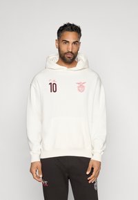 Sudadera blanca con un gran bolsillo frontal, que presenta un número 10 rojo y un emblema en el pecho. Se lleva con pantalones de chándal oscuros.