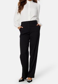 Biała bluzka z falbaniastym kołnierzykiem, czarne dopasowane spodnie oraz czarne baleriny z ostrym noskiem ozdobione kamieniami rhinestone. Gładkie, wypolerowane tekstury.