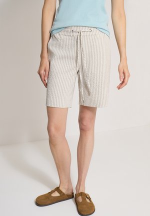 Persona con pantalones cortos beige a rayas con cordón, camiseta azul clara y sandalias marrones de ante sin cierre, de pie sobre un suelo claro.