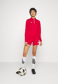 Rotes Athletic-Oberteil mit langen Ärmeln, Reißverschlusskragen und Logo, kombiniert mit passenden Shorts, weißen Socken mit schwarzen Streifen und schwarzen Fußballschuhen.