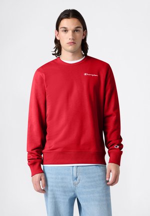 Młoda osoba z włosami do ramion, ubraną w czerwoną bluzę Champion i jasnoniebieskie dżinsy, zwrócona przodem na tle jednolitego tła.