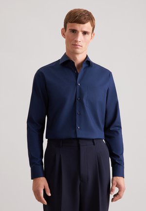 Man draagt een marineblauw hemd met knopen en een borstzak, gecombineerd met donkergekleurde, hoog gewaistte broek, staand tegen een effen achtergrond.