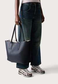 Sac en cuir bleu marine à texture avec surface texturée, poignées doubles et intérieur spacieux. Associé à un jean en denim à jambes larges et des chaussures de sport.