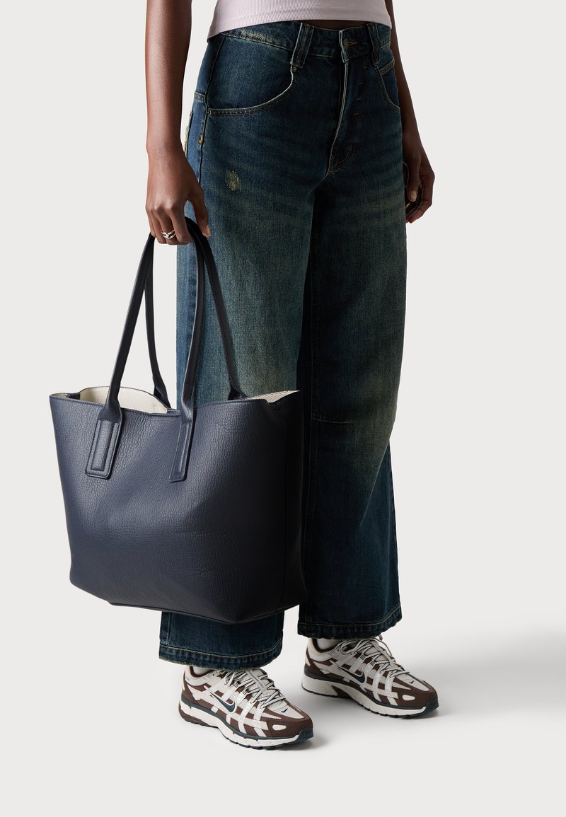Sac en cuir bleu marine à texture avec surface texturée, poignées doubles et intérieur spacieux. Associé à un jean en denim à jambes larges et des chaussures de sport.