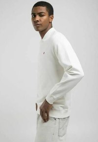 Polo shirt bianco a maniche lunghe con un piccolo logo rosso sul petto, dotato di polsini a coste e di un tessuto liscio e leggero.