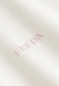 Wit stof met een gladde textuur, met het woord "FRIEDA" in lichtroze verdiept. Focus op minimalistisch ontwerp met subtiel kleurcontrast.