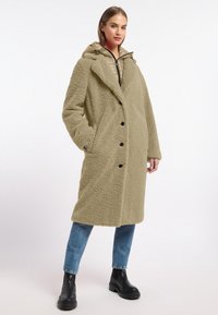 Olijfgroene jas, gemaakt van textuurstof. Beschikt over een brede kraag, grote knopen en zijzakken. Draagt over een gestreepte hoodie.