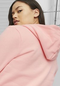 Felpa con cappuccio rosa dal tessuto morbido, caratterizzata da una vestibilità comoda e cappuccio con cordino, mostrata parzialmente con un focus sui dettagli posteriori.