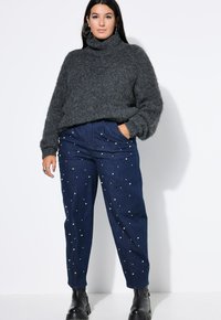 Grau-flauschiger Rollkragenpullover, kombiniert mit dunkelblauen Jeans mit weißen Perlenverzierungen, getragen mit schwarzen Knöchelstiefeln.