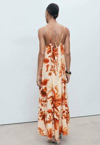Robe maxi fleurie avec un fond pêche et des motifs de feuilles orange. Elle est dotée de fines bretelles spaghetti et d'une fermeture à nouer au dos. Tissu léger.