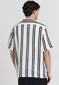 Un uomo con capelli corti e ricci indossa una maglietta bianca a maniche corte in maglia con strisce verticali nere, visto da dietro su uno sfondo neutro.