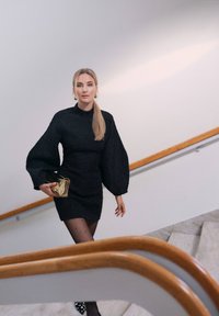 Musta teksturoitu mekko pitkillä, puffattuja hihoilla, yhdistettynä läpinäkyviin pilkullisiin sukkahousuihin ja kultaisiin pussukoihin. Taustalla on elegantti portaikko.