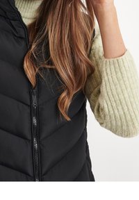 Černá prošívaná vesta s výplní a předním zipem, zkombinovaná se světle zeleným žebrovaným svetr. Materiál vypadá hladce a teple.