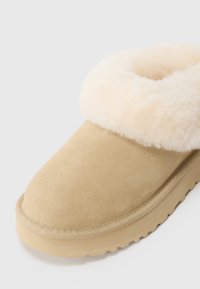 Chausson en suède beige avec une épaisse doublure en fourrure crème autour de la cheville et une semelle texturée beige.