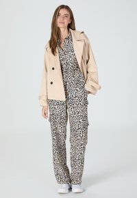 Beige trench coat over a leopard print jumpsuit, featuring black spots on a light background, paired with white sneakers. Smooth fabric texture.