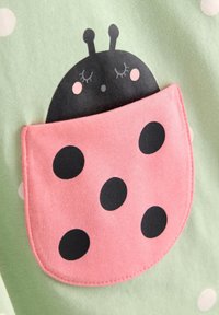 Κοιμισμένη πασχαλίτσα με ρόδινα φτερά και μαύρες κηλίδες, τυπωμένη σε τσέπη από ανοιχτό πράσινο ύφασμα με λευκές βούλες.