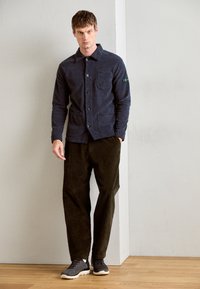 Navy button-up shirt in soft fabric with two front pockets, paired with olive corduroy pants and grey athletic shoes on a wooden floor.