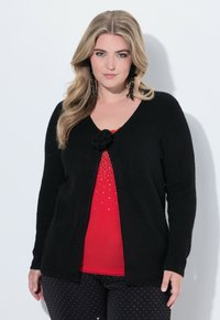 Žena s dlouhými blond vlasy nosí černý cardigan se květinovou sponou, červený top s malými kamínky a černé kalhoty s bílými tečkami.