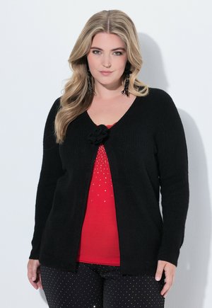 Frau mit langen blonden Haaren trägt eine schwarze Strickjacke mit Blumenverschluss, ein rotes Oberteil mit kleinen Strasssteinen und schwarze Hosen mit weißen Punkten.