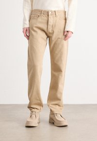 Pantaloni in cotone beige con vestibilità dritta, dotati di chiusura con bottoni e tasche frontali. Abbinati a scarpe beige che mostrano una superficie testurizzata.