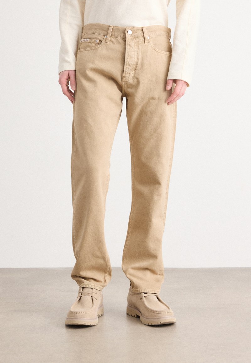 Pantaloni in cotone beige con vestibilità dritta, dotati di chiusura con bottoni e tasche frontali. Abbinati a scarpe beige che mostrano una superficie testurizzata.