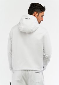 Sweat à capuche blanc avec une texture lisse, portant la marque "ICE ICEBERG" sur la capuche et le dos, associé à un pantalon de jogging assorti.
