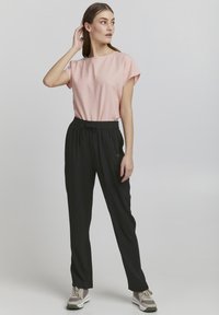 Helles rosa T-Shirt mit kurzen Ärmeln und rundem Ausschnitt, kombiniert mit schwarzen Hose mit elastischem Bund. Das Model trägt graue und weiße Sneaker.