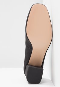 Semelle d'une chaussure noire à petit talon avec une semelle inférieure beige lisse et un talon carré noir texturé, placée contre un angle blanc.