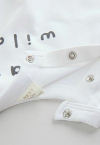 Body de bébé blanc avec fermetures à pression et texte imprimé noir. Tissu en coton doux avec une étiquette affichant des informations sur la marque.
