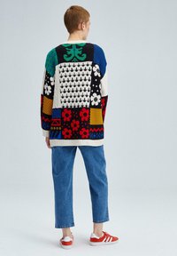 Pull en maille avec un design patchwork aux couleurs variées, présentant des motifs floraux et géométriques ; porté avec un pantalon en jean bleu et des chaussures rouges.