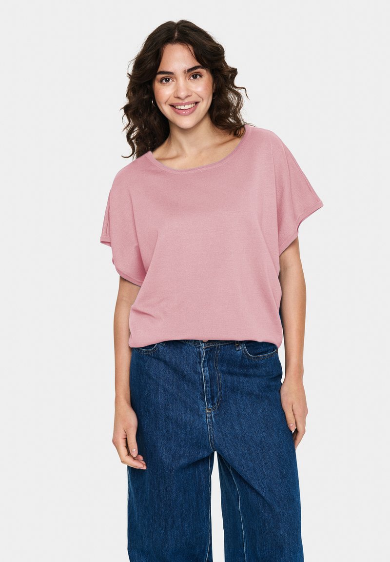Roza, ohlapna majica s okroglim izrezom in kratkimi rokavi, v kombinaciji z modrimi jeans hlačami, nihanimi visoko v pasu. Gladka tekstura tkanine.