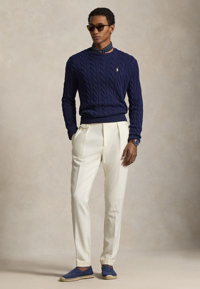 Muškarac u tamnoplavom džemperu s pletenim uzorkom, bijelim plisiranim hlačama, plavim espadrilama, tamnim sunčanim naočalama i uzorkovanom maramom oko vrata, stoji s rukama u džepovima.