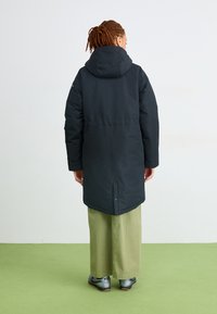 Person med fletninger iført en sort hættejakke, lysegrønne brede bukser og sølvfarvede sneakers, der står på grøn gulv og har ryggen til.