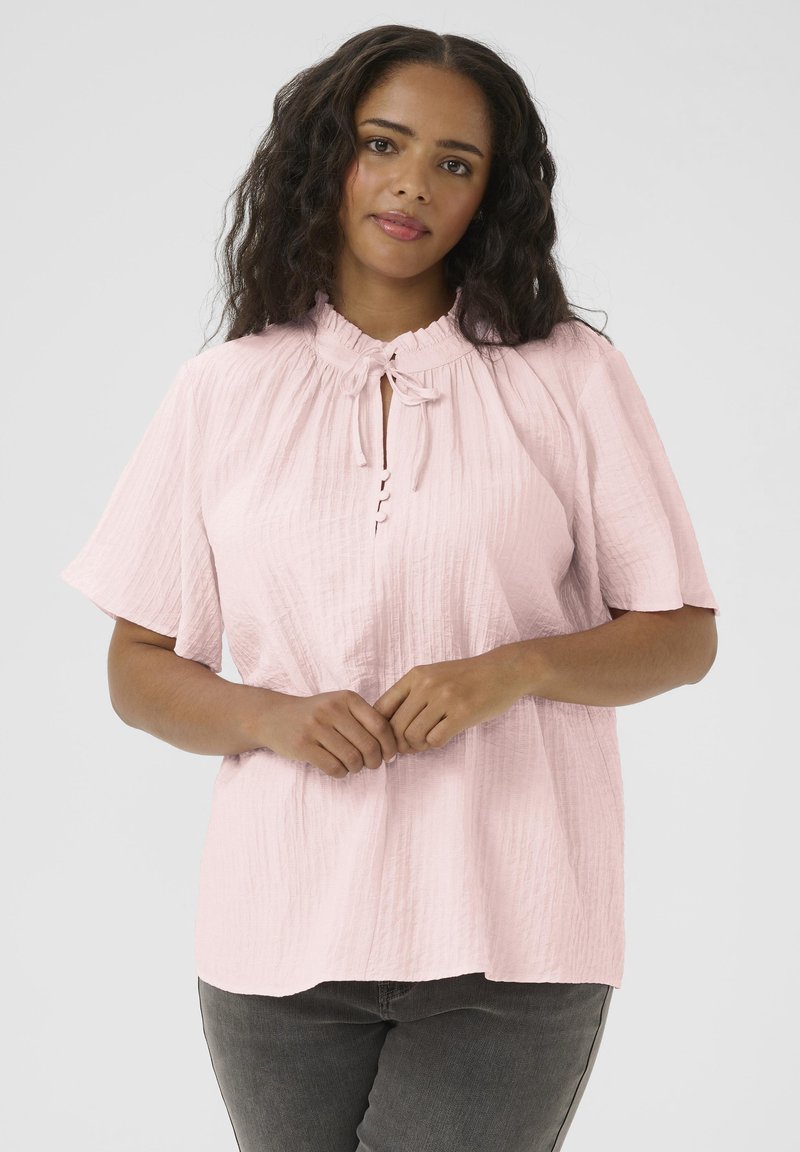 Blouse rose clair en tissu texturé, présentant un col haut avec un lien et une patte de boutonnage sur le devant. Manches courtes.