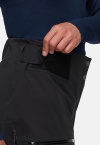 Μαύρο παντελόνι με κλείσιμο από βέλκρο και κουμπί, διαθέτοντας φερμουάρ στις τσέπες και λεία υφή. Ένα χέρι ρυθμίζει την περιοχή της μέσης.