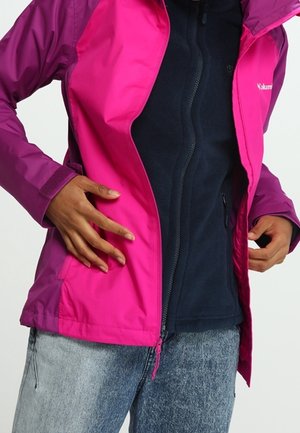 Person trägt eine leuchtend pinke Jacke über einem schwarzen Zip-Up-Top und hellblauen Jeans, mit Händen, die die Seiten der Jacke anpassen.