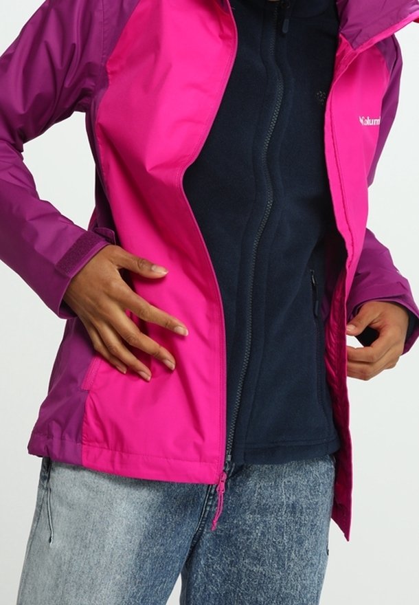Personne portant une veste rose vif sur un haut noir zippé et un jean bleu clair, les mains ajustant les côtés de la veste.