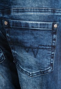 Džínové kalhoty s tmavě modrým praním, s kapsou na zadní straně zdobenou dekorativním prošíváním a kovovým knoflíkem. Texturovaný povrch, neformální design.