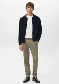 Jonge man met een zwart gebreid vest met knopen over een wit overhemd, beige ribfluwelen broek en donkerbruine leren laarzen, staand met handen in de zakken.