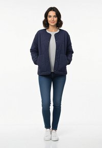 Frau steht mit Händen in den Taschen, trägt eine marineblaue Steppjacke, ein graues Shirt, dunkelblaue Skinny-Jeans und weiße Sneakers vor einem weißen Hintergrund.